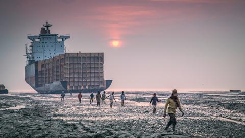 Ein Schiff liegt am Strand in Chittagong, Bangladesh und wird von Arbeitern auseinandergenommen. Vor dem Sonnenuntergang gehen Arbeiter in Richtung der Kamera.