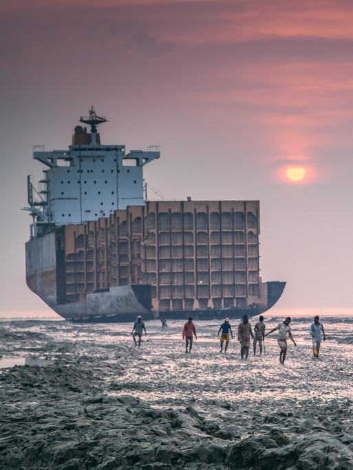 Ein Schiff liegt am Strand in Chittagong, Bangladesh und wird von Arbeitern auseinandergenommen. Vor dem Sonnenuntergang gehen Arbeiter in Richtung der Kamera.