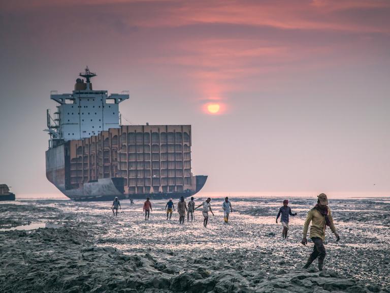 Ein Schiff liegt am Strand in Chittagong, Bangladesh und wird von Arbeitern auseinandergenommen. Vor dem Sonnenuntergang gehen Arbeiter in Richtung der Kamera.