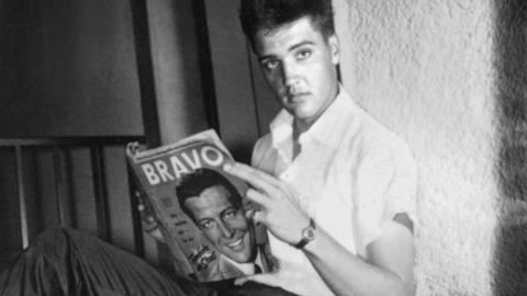 Der US-Sänger Elvis Presley in seinem möbilierten Zimmer in Bad Nauheim während der Stationierung als US-Soldat. Er blättert in einer Ausgabe der Jugendzeitschrift "Bravo". Das Foto ist schwarz-weiß. 