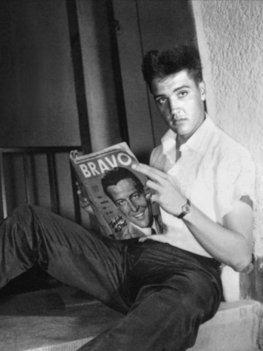 Der US-Sänger Elvis Presley in seinem möbilierten Zimmer in Bad Nauheim während der Stationierung als US-Soldat. Er blättert in einer Ausgabe der Jugendzeitschrift "Bravo". Das Foto ist schwarz-weiß. 