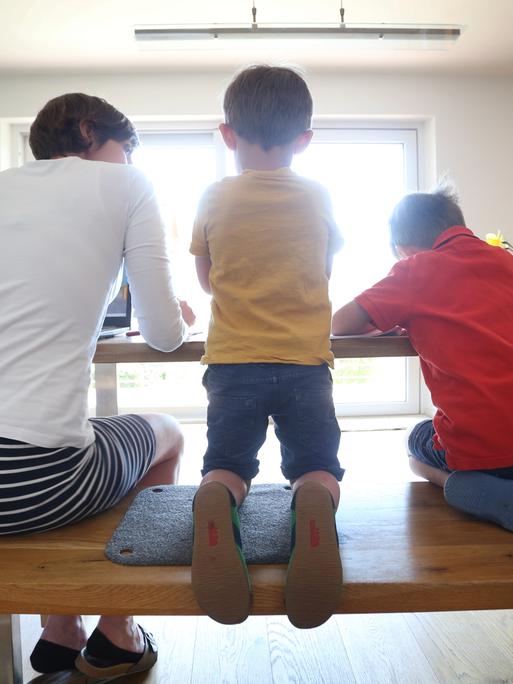 Eine Mutter und zwei Kinder sitzen mit dem Rücken zum Betrachter an einem Küchentisch.