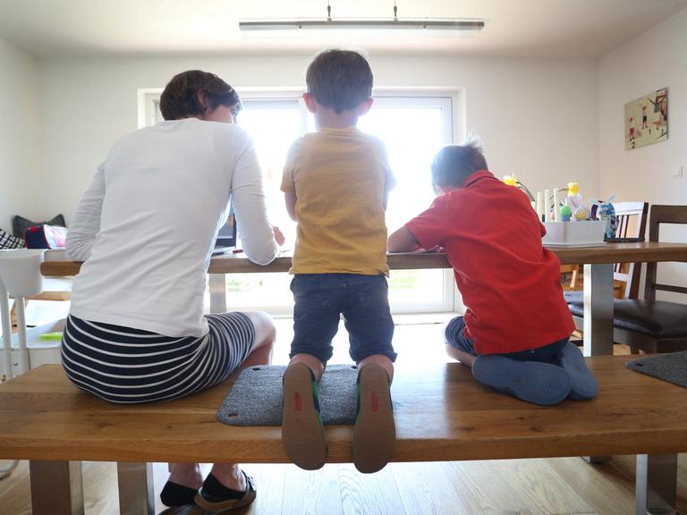 Eine Mutter und zwei Kinder sitzen mit dem Rücken zum Betrachter an einem Küchentisch.