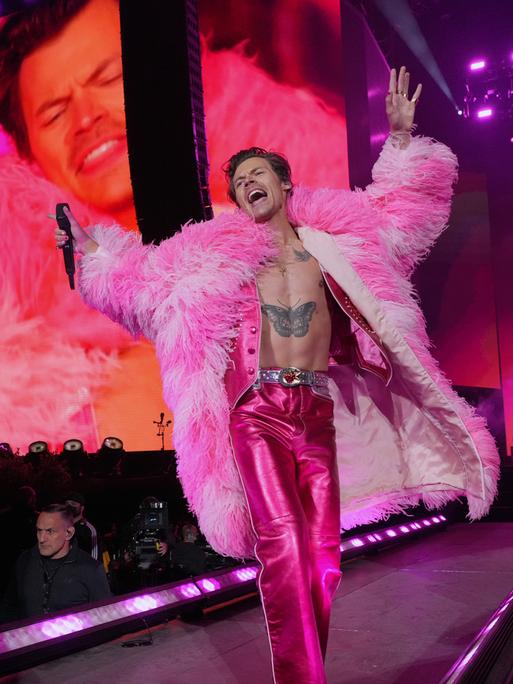 Harry Styles performt auf der Bühne. Er trägt einen Mantel aus pinkfarbenen Federn.