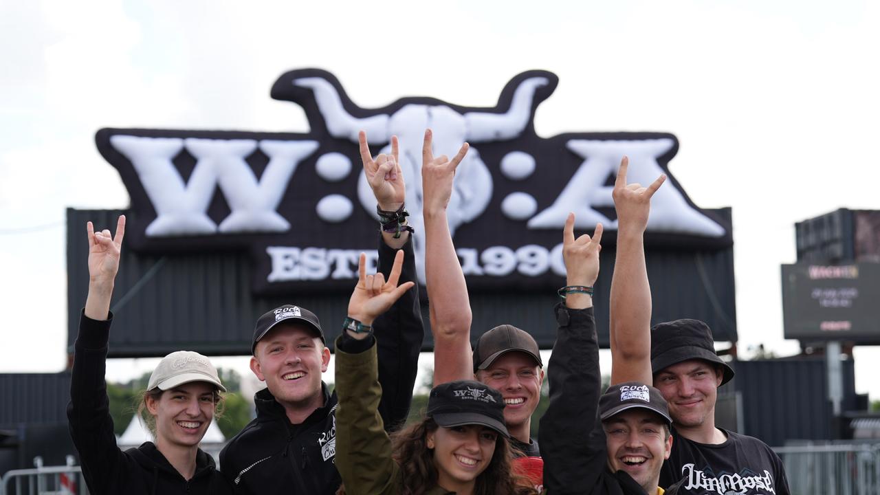 Mehrere Menschen stehen vor dem Eingangsschild des Wacken-Festivals und machen einen Heavy-Metal-Gruß.