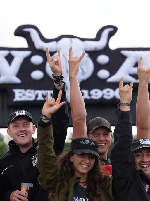 Mehrere Menschen stehen vor dem Eingangsschild des Wacken-Festivals und machen einen Heavy-Metal-Gruß.