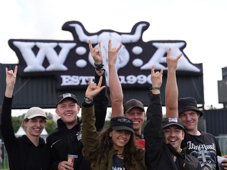 Mehrere Menschen stehen vor dem Eingangsschild des Wacken-Festivals und machen einen Heavy-Metal-Gruß.