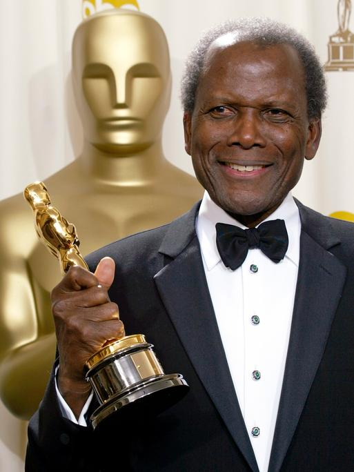Der schwarze Schauspieler Sidney Poitier steht neben einer großen Oscar-Statue und hält einen Oscar in der Hand