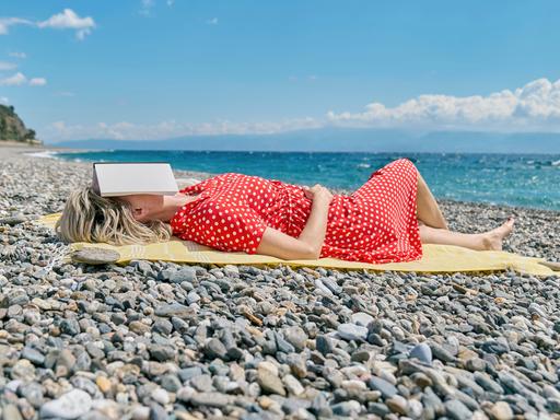 Eine Frau liegt in einem roten Kleid mit weißen Punkten an einem steinigen Strand. Auf ihrem Gesicht liegt ein geöffnetes Buch.