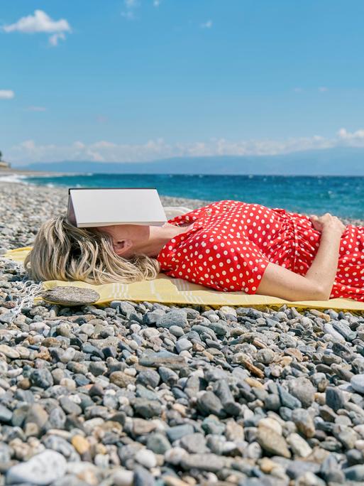Eine Frau liegt in einem roten Kleid mit weißen Punkten an einem steinigen Strand. Auf ihrem Gesicht liegt ein geöffntetes Buch.