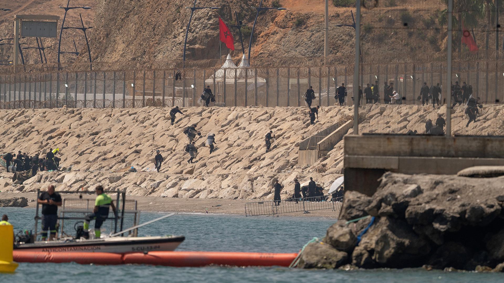 Marokkanische Soldaten stehen am Grenzzaun zwischen Marokko und der spanischen Exklave Ceuta in Nordafrika. Einige Menschen klettern vom Meer aus in Richtung Zaun. 