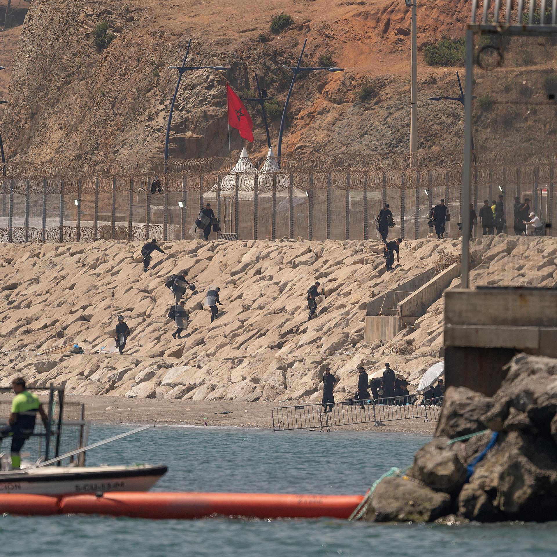 Marokkanische Soldaten stehen am Grenzzaun zwischen Marokko und der spanischen Exklave Ceuta in Nordafrika. Einige Menschen klettern vom Meer aus in Richtung Zaun.  