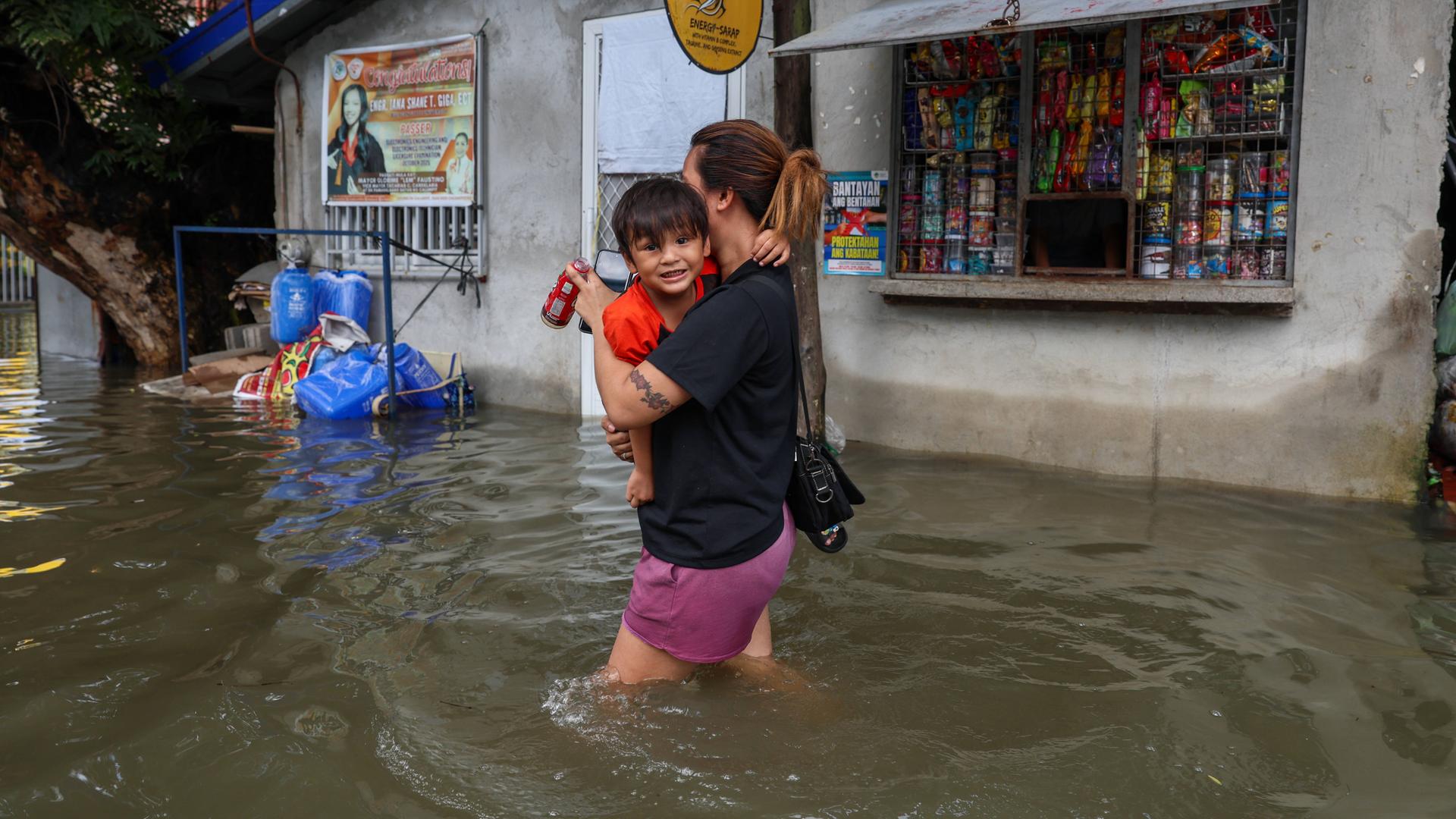 Eine Frau trägt ihr Kleinkind auf dem Arm und watet durch die überschwemmte Straße. Das Wasser reicht ihr bis zu den Oberschenkeln. Im Hintergrund ist ein geöffneter Kiosk.