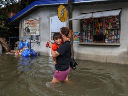 Eine Frau trägt ihr Kleinkind auf dem Arm und watet durch die überschwemmte Straße. Das Wasser reicht ihr bis zu den Oberschenkeln. Im Hintergrund ist ein geöffneter Kiosk.