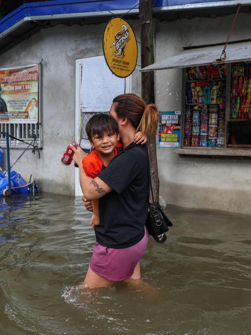 Eine Frau trägt ihr Kleinkind auf dem Arm und watet durch die überschwemmte Straße. Das Wasser reicht ihr bis zu den Oberschenkeln. Im Hintergrund ist ein geöffneter Kiosk.