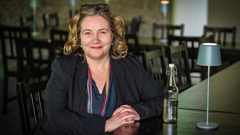 Eine Frau sitzt in die Kamera lächelnd in einem leeren Restaurant. Sie hat ihre Brille in die Haare geschoben und trägt über ihrem dunklen Outfit ein dünnes Tuch, das viele bunte Streifen zeigt.