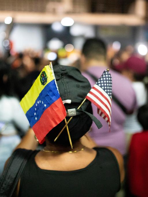 Fähnchen von Venezuela und den USA stecken in einer Kappe einer Frau inmitten einer Menschenmenge