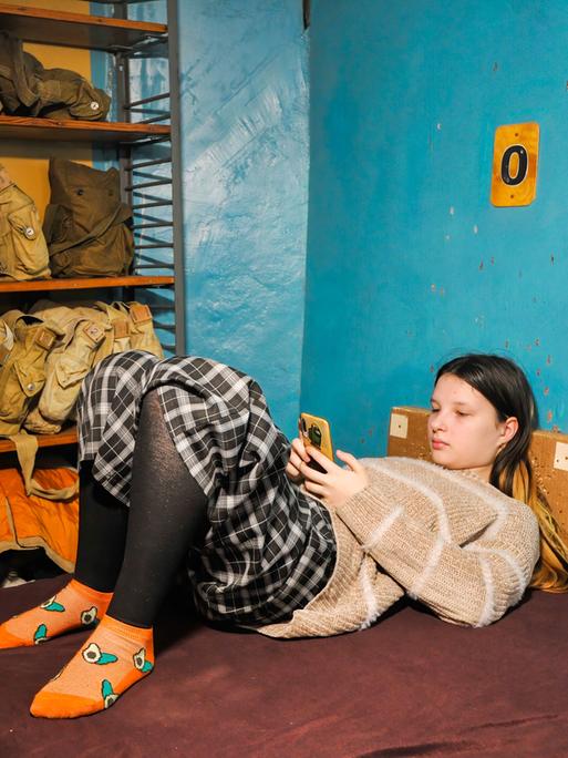 Eine Jugendliche liegt auf einer Liege in einem Luftschutzbunker und schaut auf ihr Smartphone.