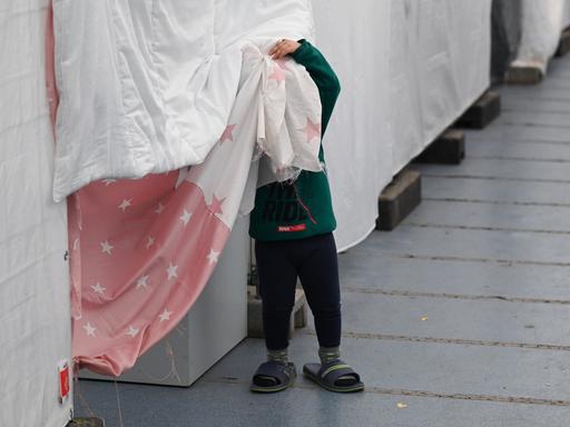 Ein Flüchtlingskind steht in einem Zelt der Flüchtlingsunterkunft vor einer Wohnparzelle und verdeckt sein Gesicht.