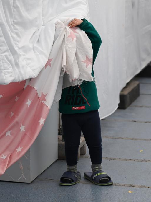 Ein Flüchtlingskind steht in einem Zelt der Flüchtlingsunterkunft vor einer Wohnparzelle und verdeckt sein Gesicht.