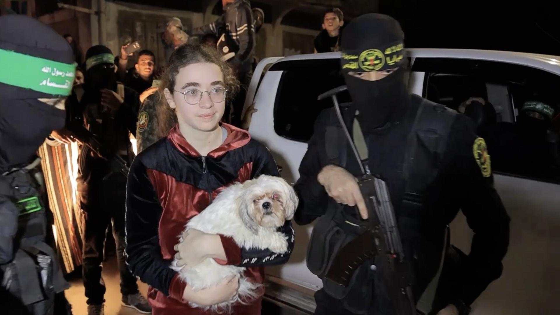Ein Junge mit einem weißen Hund auf dem Arm wird von zwei schwarz vermummten Hamas- Mitgliedern begleitet.