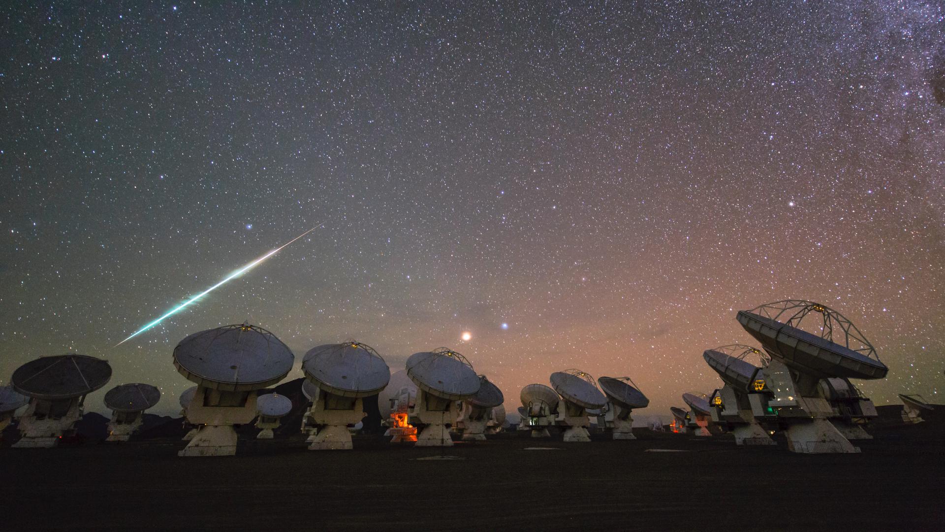 Meteorschweif am Sternenhimmel vor der Silhouette einer Teleskopanlage mit großer Radarschüssel.