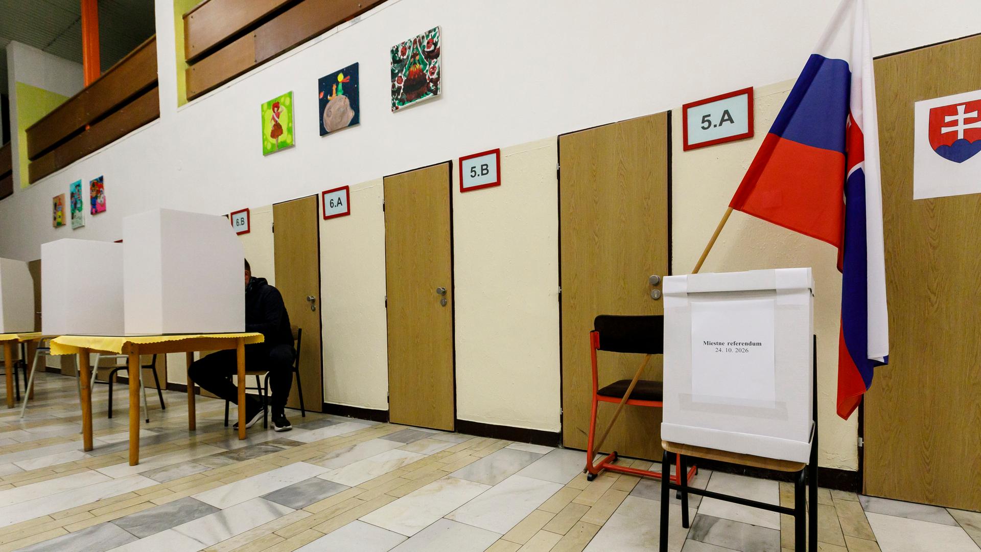 Blick in das Stimmlokal in Bratislava, Slowakei: Mehrere viereckige Sichtschutze aus Pappe stehen auf Tischen, hinter einem gibt gerade ein Mann seine Stimme ab. Am rechten Bildrand auf einem Tisch die Wahlurne, dahinter hängt die slowakische Flagge. 