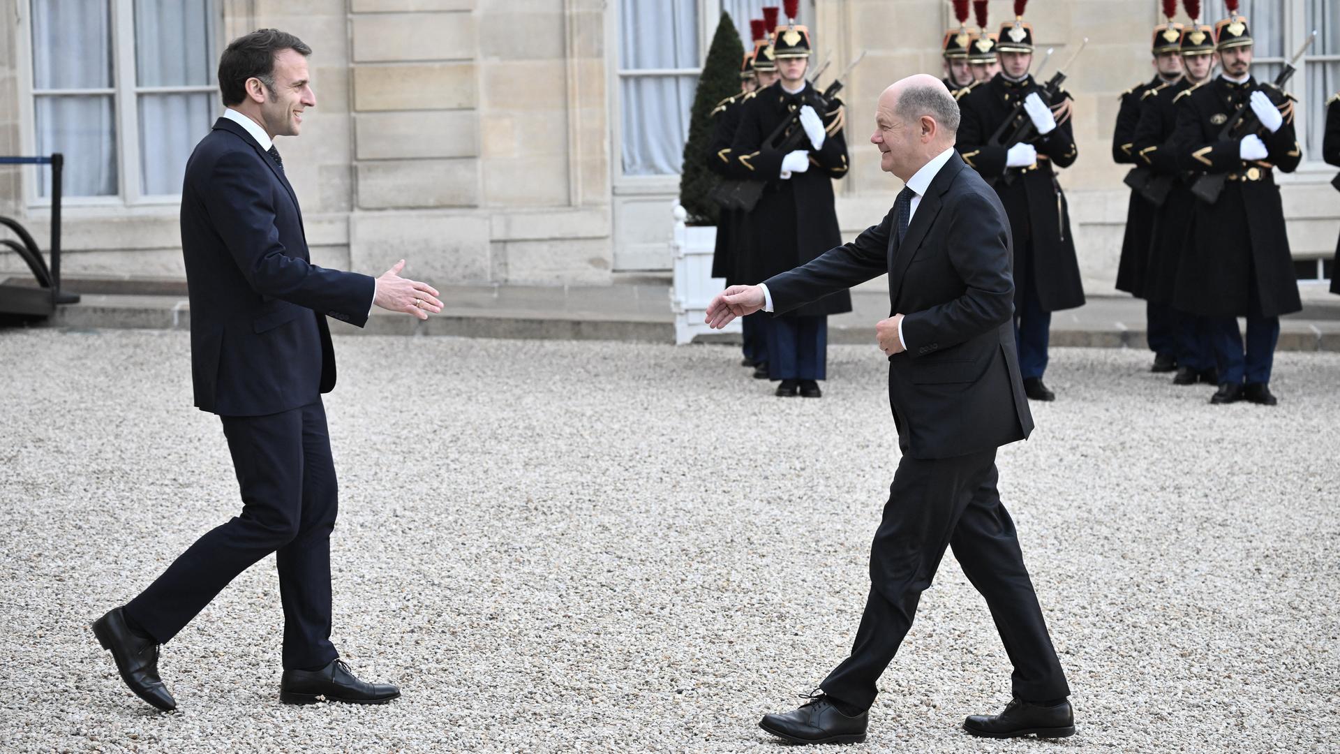 Emmanuel Macron geht mit ausgestreckter Hand auf Olaf Scholz zu. Im Hintergrund stehen französische Soldaten.