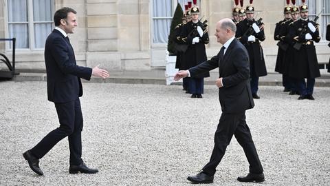 Emmanuel Macron geht mit ausgestreckter Hand auf Olaf Scholz zu. Im Hintergrund stehen französische Soldaten.