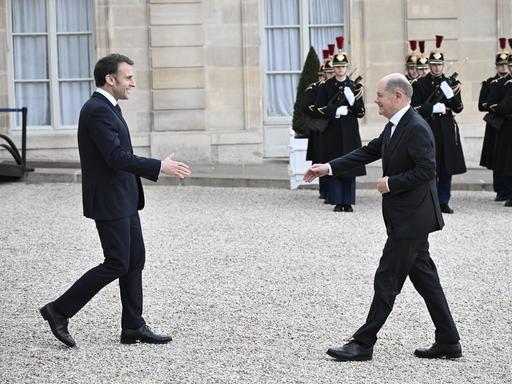 Emmanuel Macron geht mit ausgestreckter Hand auf Olaf Scholz zu. Im Hintergrund stehen französische Soldaten.