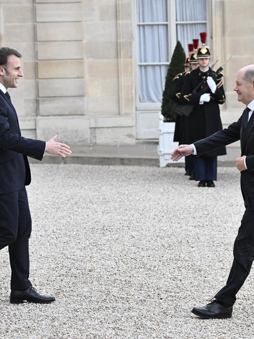 Emmanuel Macron geht mit ausgestreckter Hand auf Olaf Scholz zu. Im Hintergrund stehen französische Soldaten.