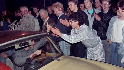 Ein Mann fährt in seinem Trabant-Automobil  in der Nacht vom 9. auf den 10. November 1989 über den Grenzübergang Bornholmer Brücke in Berlin. Andere Menschen, im Vordergrund ein Mann mit silberfarbenen Anzug, applaudieren.