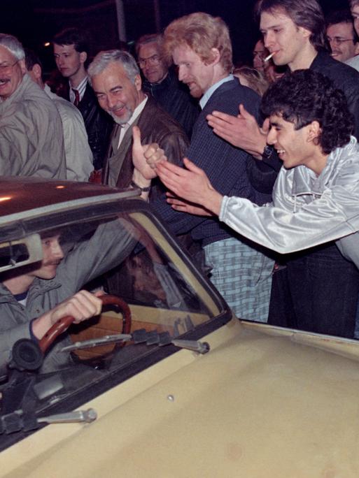 Ein Mann fährt in seinem Trabant-Automobil  in der Nacht vom 9. auf den 10. November 1989 über den Grenzübergang Bornholmer Brücke in Berlin. Andere Menschen, im Vordergrund ein Mann mit silberfarbenen Anzug, applaudieren.