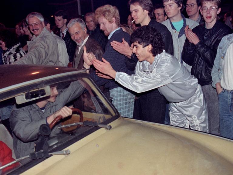 Ein Mann fährt in seinem Trabant-Automobil  in der Nacht vom 9. auf den 10. November 1989 über den Grenzübergang Bornholmer Brücke in Berlin. Andere Menschen, im Vordergrund ein Mann mit silberfarbenen Anzug, applaudieren.