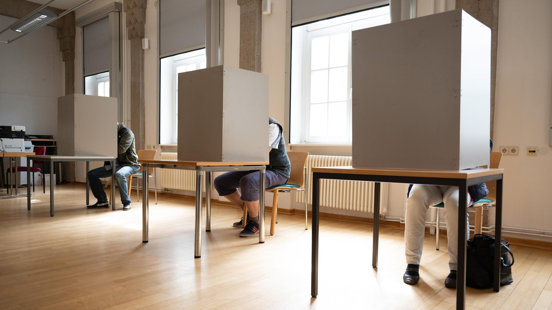 Drei Menschen sitzen in einem Brief-Wahl-Büro hinter Aufstell-Wänden, um ihre Stimm-Zettel auszufüllen.