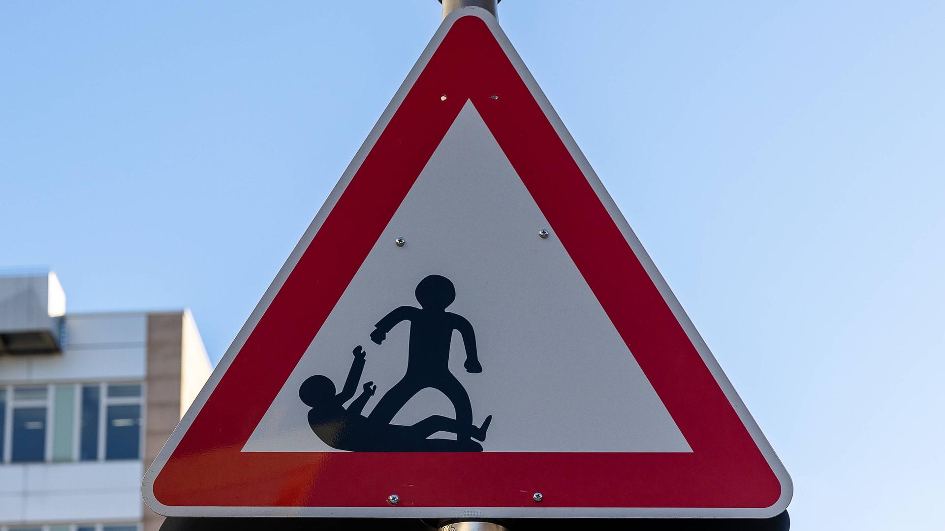 Dreieckiges Warnschild mit zwei schwarzen Figuren, von denen eine aggressiv erscheint und die andere am Boden liegt. Das rechteckige Schild darunter trägt die Aufschrift "SCHAU HIN".