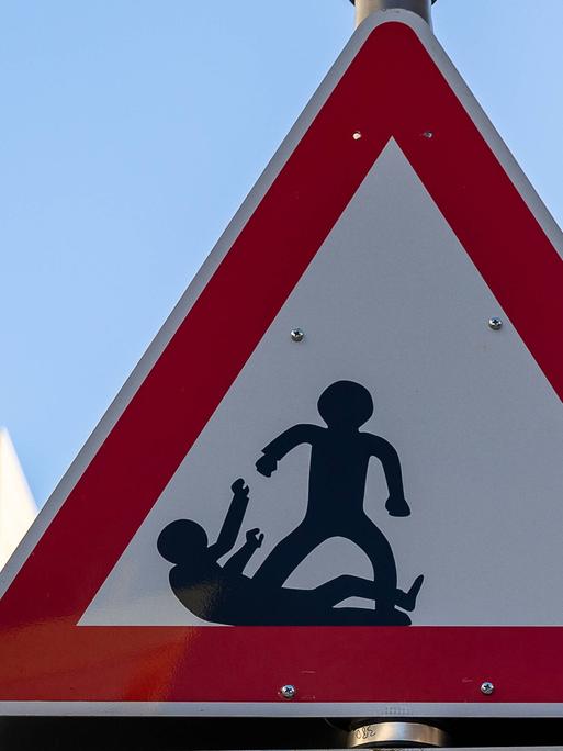 Dreieckiges Warnschild mit zwei schwarzen Figuren, von denen eine aggressiv erscheint und die andere am Boden liegt. Das rechteckige Schild darunter trägt die Aufschrift "SCHAU HIN".