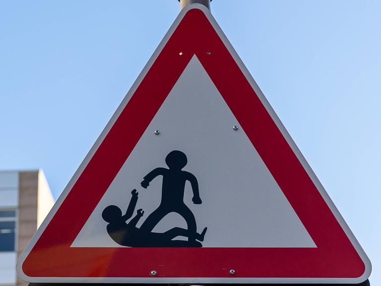 Dreieckiges Warnschild mit zwei schwarzen Figuren, von denen eine aggressiv erscheint und die andere am Boden liegt. Das rechteckige Schild darunter trägt die Aufschrift "SCHAU HIN".