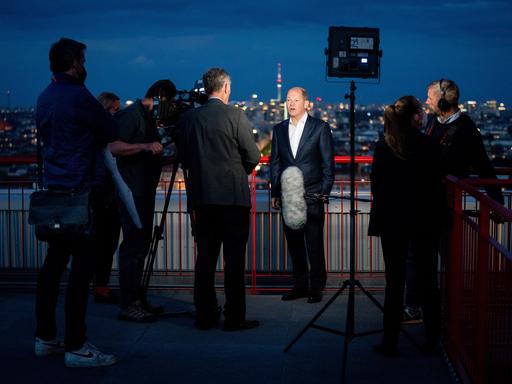 Olaf Scholz (SPD) steht 2021 vor der abendlichen Berliner Skyline auf einer Dachterasse und gibt dem Dokumentarfilmer Stephan Lamby (3.vl) ein Interview.