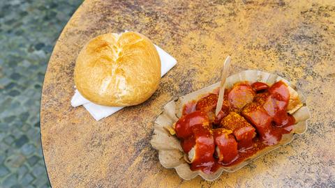 EIne Currywurst und ein Brötchen liegen auf einem Tisch vor Konnopkes Imbiss, dem Kult-Imbissstand im Berliner Stadtteil Prenzlauer Berg. Konnopke gilt als erster Imbissstand in Ost-Berlin, bei dem im Jahr 1960 die Currywurst eingeführt wurde.