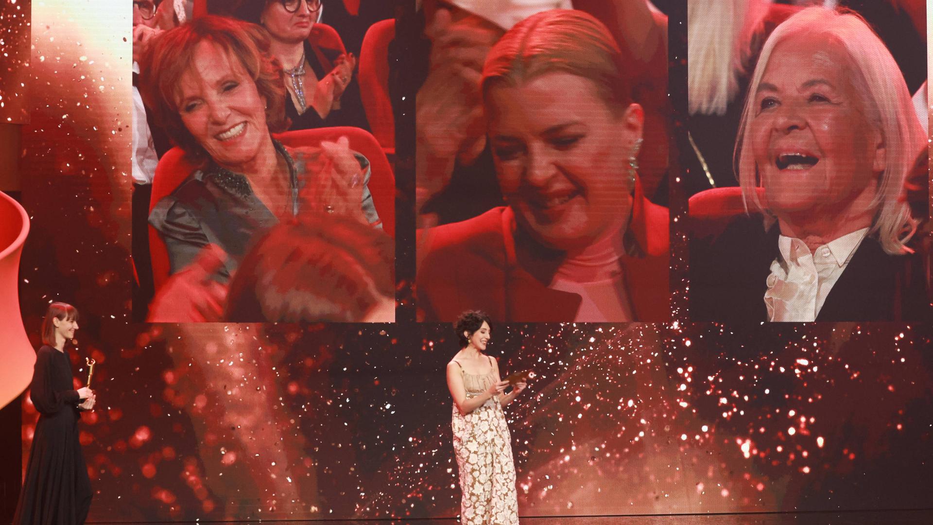 Deutscher Filmpreis 2023: Die Auszeichnung für die beste weibliche Nebenrolle geht an Jördis Triebel. Sie ist auf einer Leinwand hinter der Bühne zu sehen. Auf der Bühne steht die Laudatorin. Deutscher Filmpreis 2023: Die Auszeichnung für die beste weibliche Nebenrolle geht an Jördis Triebel. Sie ist auf einer Leinwand hinter der Bühne zu sehen. Auf der Bühne steht die Laudatorin.
