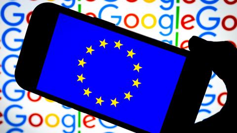 Ein Handy zeigt die EU-Sterne und wird vor einen Hintergrund gehalten, vor dem mehrfach Google steht. 