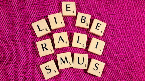 Scrabble-Buchstaben auf pinkem Untergrund bilden das Wort Liberalismus - zwischen dem I und dem B scheint sich ein E dazwischendrängeln zu wollen, das Wort Liebe wird angedeutet.