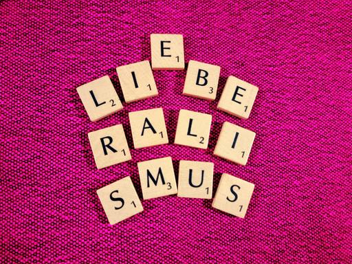 Scrabble-Buchstaben auf pinkem Untergrund bilden das Wort Liberalismus - zwischen dem I und dem B scheint sich ein E dazwischendrängeln zu wollen, das Wort Liebe wird angedeutet.