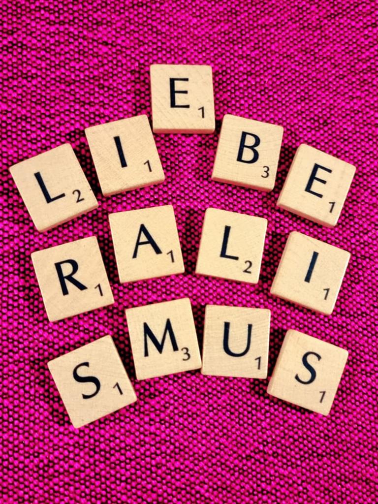 Scrabble-Buchstaben auf pinkem Untergrund bilden das Wort Liberalismus - zwischen dem I und dem B scheint sich ein E dazwischendrängeln zu wollen, das Wort Liebe wird angedeutet.