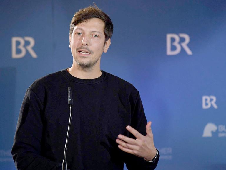 Thilo Mischke steht vor einem Fotohintergrund des BR und spricht am Mikrofon Thilo Mischke steht vor einem Fotohintergrund des BR und spricht am Mikrofon