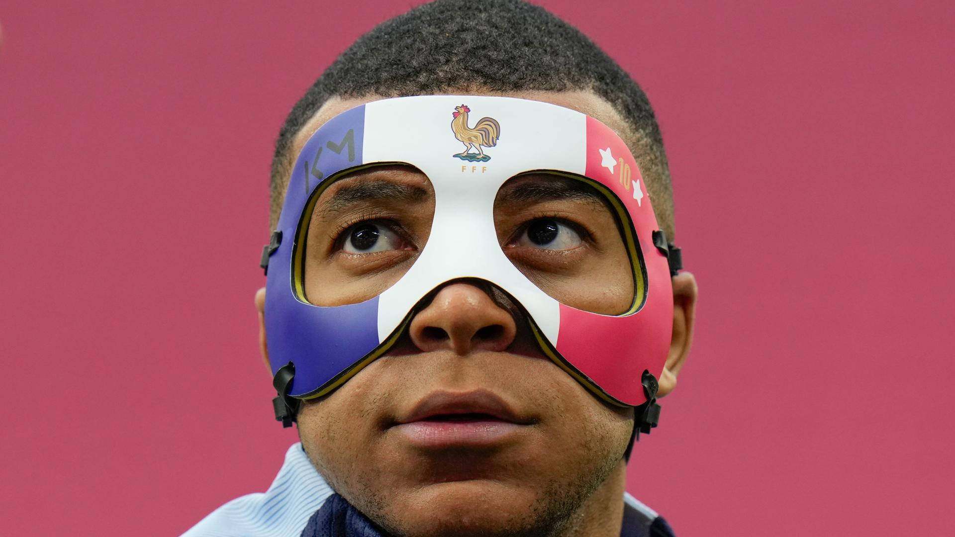Kylian Mbappé mit einer Gesichtsmaske, die die Augen einrahmt und über den oberen Teil der Nase ragt. Die Maske ist in den Farben der französischen Flagge gestaltet. Dazu trägt sie Mbappés Logo KY, das Logo des französischen Fußballverbands in Form eines Hahns und Mbappés Rückennummer 10 zwischen Sternen.