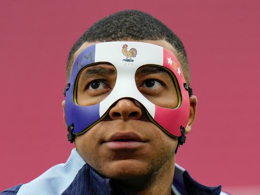 Kylian Mbappé mit einer Gesichtsmaske, die die Augen einrahmt und über den oberen Teil der Nase ragt. Die Maske ist in den Farben der französischen Flagge gestaltet. Dazu trägt sie Mbappés Logo KY, das Logo des französischen Fußballverbands in Form eines Hahns und Mbappés Rückennummer 10 zwischen Sternen.