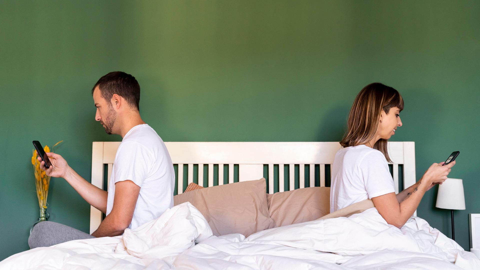 2 Erwachsene sitzen auf dem Bett. Beide gucken auf das Handy.