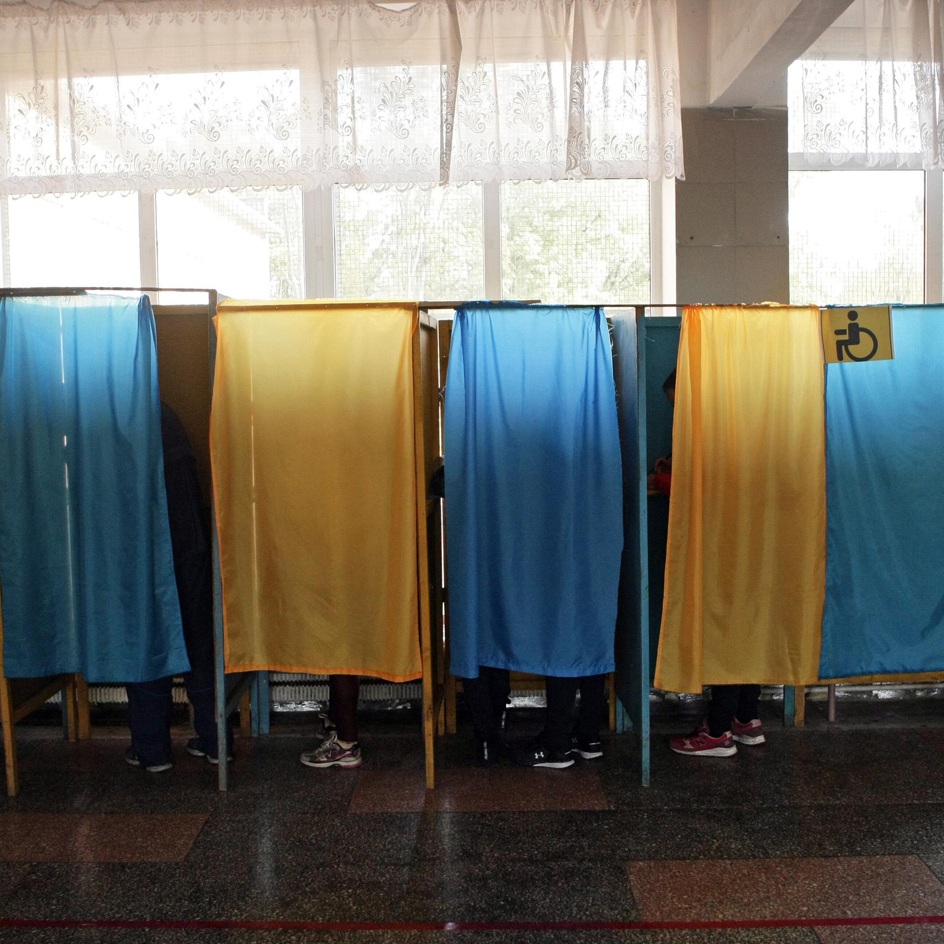 Blaue und gelbe Vorhänge an Wahlkabinen vor einem Fenster.